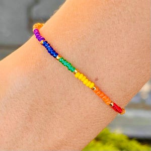 May include: A rainbow beaded bracelet with gold accents. The bracelet is made with small, colorful beads in the order of red, orange, yellow, green, blue, and purple.