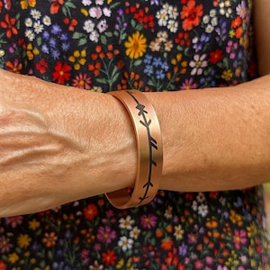Peut inclure: Un bracelet manchette de couleur cuivre avec un motif de flèches noires. Le bracelet est porté au poignet, sur un fond floral aux couleurs variées.