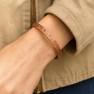 May include: A copper-colored cuff bracelet with a hammered texture. The bracelet is a simple, open design, and is worn on a wrist. The background includes a tan jacket.