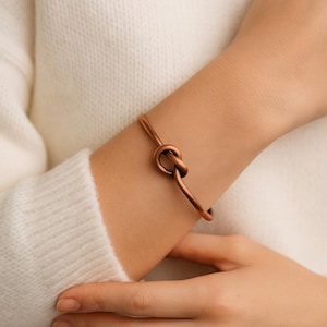May include: A copper-colored bracelet with a knot design. The bracelet is a simple, elegant cuff style, with a knot as the focal point. The metal has a warm, reddish-brown hue. The bracelet is worn on a wrist.