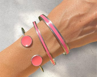 May include: Three silver bracelets with pink enamel accents. The bracelets are stacked on a person's wrist. The bracelets have a simple, modern design.