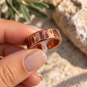 Peut inclure: Une bague en cuivre avec une texture martelée et un design ouvert. Le métal présente une finition métallique brun rougeâtre chaud avec des indentations visibles sur la surface extérieure, créant un aspect réfléchissant et irrégulier.
