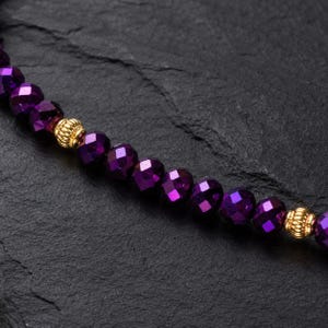 May include: A close-up of a beaded necklace featuring faceted, deep purple beads. The necklace has gold-coloured, textured accent beads. The beads are strung together, creating a shimmering effect.