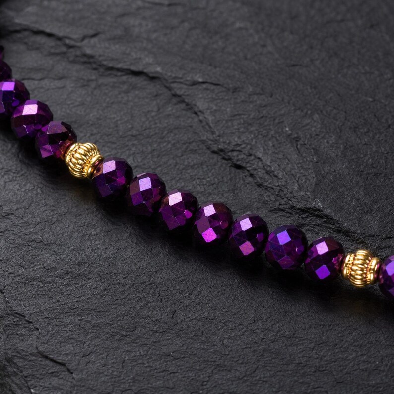 May include: A close-up of a beaded necklace featuring faceted, deep purple beads. The necklace has gold-coloured, textured accent beads. The beads are strung together, creating a shimmering effect.