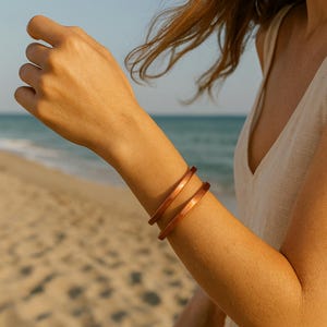 May include: Two copper-colored bangles are worn on an arm, with a beach and ocean in the background. The bangles are a warm, reddish-brown hue, and appear to be made of metal. The arm is fair-skinned, and the person is wearing a light-colored top.