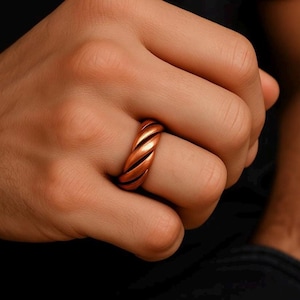 May include: A copper-coloured ring with a twisted design. The ring is worn on a finger, showcasing its spiral pattern and metallic sheen. The ring's design features a series of parallel grooves, creating a textured surface.