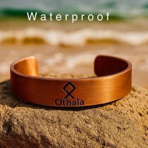 May include: A copper-colored cuff bracelet with the word "Waterproof" above it. The bracelet has the word "Othala" and a symbol engraved on it. The bracelet is resting on a rock.