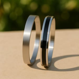 Könnte beinhalten: Zwei silberfarbene Ringe. Ein Ring ist silberfarben, der andere hat einen schwarzen Streifen in der Mitte. Die Ringe werden auf einer hellbraunen Oberfläche mit verschwommenem grünem Hintergrund gezeigt.
