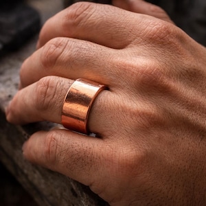 Op de afbeelding: Een brede, koperkleurige ring met een glad, gepolijst oppervlak. De ring is een massieve band, met een minimalistisch ontwerp. De warme, roodbruine tint van het metaal is prominent aanwezig en reflecteert licht. Dit sieraad is een statement-accessoire.