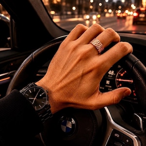 Könnte beinhalten: Eine Hand mit einem strukturierten roségoldenen Ring hält ein Lenkrad. Eine schwarze Uhr mit dunklem Zifferblatt ist am Handgelenk zu sehen. Der Hintergrund zeigt eine verschwommene Stadtstraße bei Nacht.
