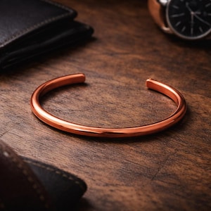 May include: A copper-colored cuff bracelet with a rounded design. The open-ended bracelet is displayed on a wooden surface, with a wallet and watch in the background. The bracelet is a simple, minimalist design.