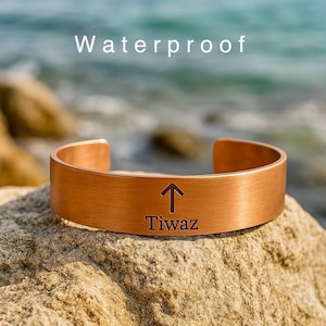 Könnte beinhalten: Ein kupferfarbenes Armreif-Armband mit dem Wort "Tiwaz" und einem nach oben zeigenden Pfeil auf der Vorderseite. Das Wort "Waterproof" steht über dem Armband. Das Armband liegt auf einem Felsen in der Nähe des Ozeans.
