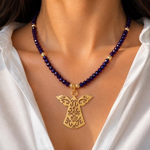 May include: A gold angel pendant necklace with a dark purple beaded strand. The necklace features gold accents and is worn against a white shirt. The angel pendant has an intricate, openwork design.