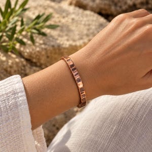 May include: A copper cuff bracelet featuring a distinct hammered texture along its entire outer surface. The metal has a warm, reddish-brown metallic finish with reflective highlights across the indented, rhythmic pattern of the band.