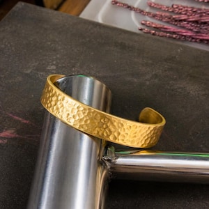 May include: A gold-coloured cuff bracelet with a hammered texture. The open-ended bracelet is resting on a silver metal object. The bracelet has a textured surface and a curved shape.