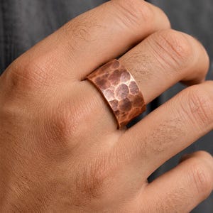 May include: A wide copper ring with a hammered texture. The ring has a band-like design and a warm, reddish-brown color. The ring is worn on a finger.