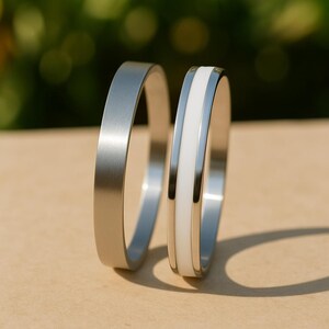 May include: Two silver rings, one solid and the other with a white enamel inlay. The rings are displayed on a light brown surface, with a blurred green background. The rings are made of a shiny metal.