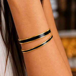 May include: Two gold bangles with black enamel detailing are shown on an arm. The bangles have a modern design, with a thin gold border framing the black enamel. The jewellery is displayed on a person's arm.