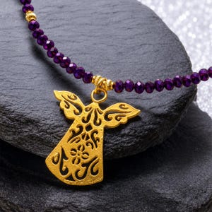 May include: A gold angel pendant necklace with a purple beaded chain. The angel pendant features detailed cut-out patterns. The necklace is presented on a dark grey, textured surface.