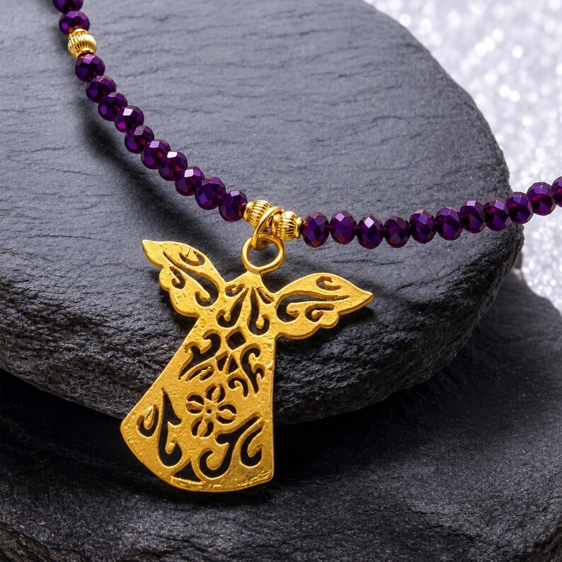 May include: A gold angel pendant necklace with a purple beaded chain. The angel pendant features detailed cut-out patterns. The necklace is presented on a dark grey, textured surface.