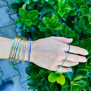 May include: A set of five thin gold-tone bangles and four matching rings, each featuring a central inlay of vibrant glittery enamel in green, red, blue, yellow, and purple, worn together on a wrist and fingers against a background of green leaves.