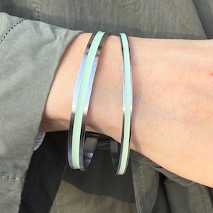 Pode incluir: Duas pulseiras prateadas com uma fina faixa verde em cada uma. As pulseiras estão empilhadas no pulso de uma pessoa.