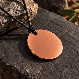Puede incluir: Un colgante circular de cobre con un acabado mate y liso, suspendido de un cordón negro. El colgante presenta un único orificio en la parte superior para pasar el cordón, creando un diseño sencillo que resalta la superficie metálica.