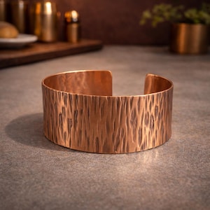 Può includere: Un ampio bracciale a fascia realizzato in rame lucido caratterizzato da una texture martellata verticale su tutta la superficie esterna. Il metallo presenta una calda tonalità bruno-rossastra con aree più scure all'interno dei solchi del bracciale.