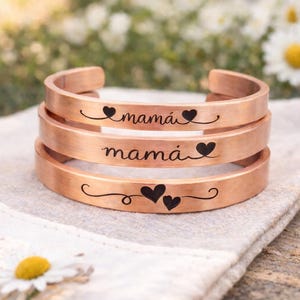 May include: Three copper-colored cuff bracelets stacked on top of each other. Each bracelet has the word "mamá" engraved in black script, with heart accents. The bracelets are resting on a light-colored cloth.