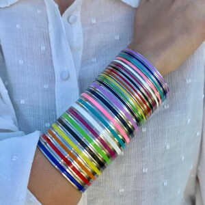 Puede incluir: Una pila de pulseras de esmalte de colores en la muñeca de una persona. Las pulseras son de diferentes colores, incluyendo rojo, naranja, amarillo, verde, azul, morado y rosa. Están apiladas unas sobre otras, creando un efecto arcoíris.