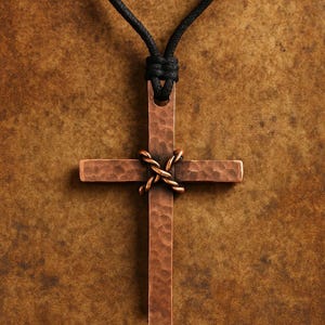 Peut inclure: Pendentif en forme de croix de couleur cuivre, avec une surface texturée, suspendu à un cordon noir. La croix est ornée d'un accent en cuivre tressé à l'intersection des bras. Le fond est marron moucheté.