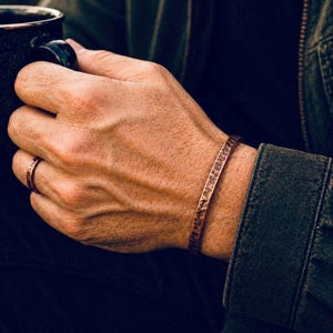 Op de afbeelding: Een koperkleurige armband en ring set. De armband is een gehamerde manchetstijl en de ring is een eenvoudige band. De sieraden worden gedragen aan de hand van een persoon, met een donkere achtergrond.