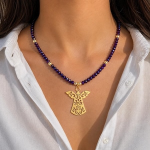 May include: A gold angel pendant necklace with a purple beaded chain. The pendant features a detailed, openwork design. The necklace is worn against a white shirt.
