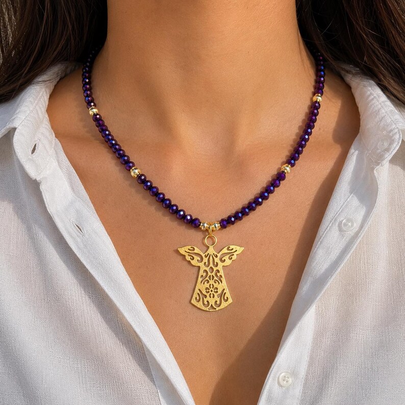 May include: A gold angel pendant necklace with a purple beaded chain. The pendant features a detailed, openwork design. The necklace is worn against a white shirt.