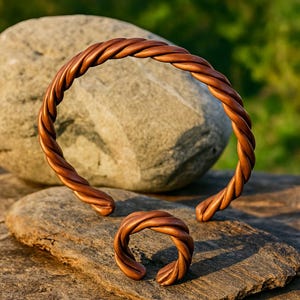 Op de afbeelding: Een set handgemaakte koperen sieraden, waaronder een armband en ring met een gedraaid touwontwerp. De warme bruine tinten van het metaal contrasteren met de natuurlijke stenen achtergrond. De ring is maat 17 (NL).