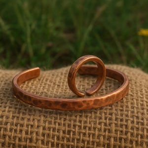 May include: A copper cuff bracelet and a matching copper ring, both featuring a hammered, textured surface. The bracelet is an open-ended circular band, and the ring is a smaller, open-ended band, both displaying a warm, metallic reddish-brown finish.