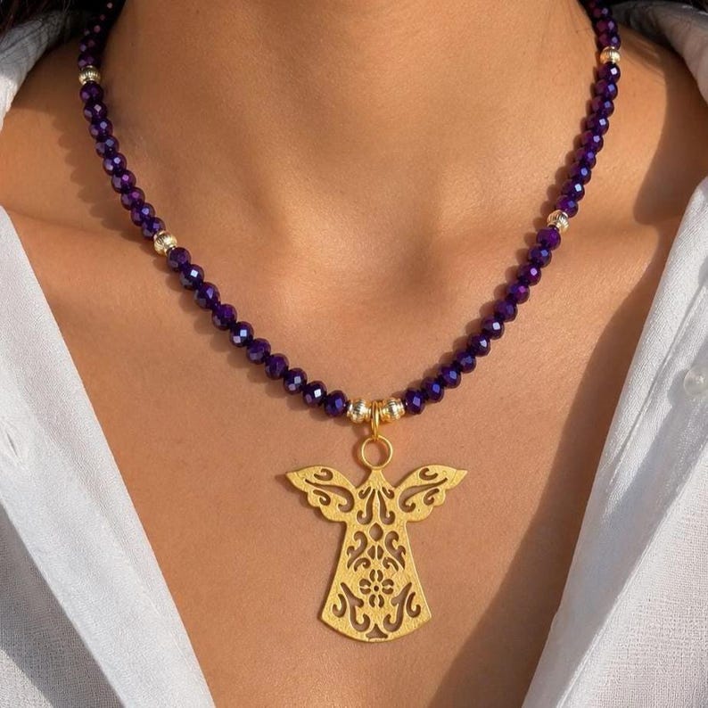 May include: A gold angel pendant necklace with a beaded purple amethyst chain. The pendant features an intricate openwork design. The necklace is worn against a white shirt.