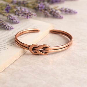 Op de afbeelding: Een koperkleurige armband met een knoopontwerp. De open manchetarmband is gemaakt van twee parallelle banden die samenkomen bij de knoop. De armband ligt op een boek met lavendel in de achtergrond.
