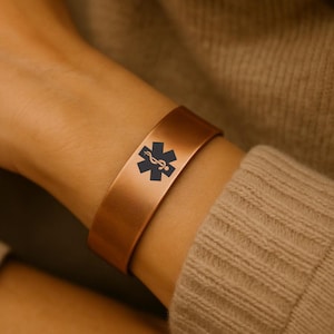 May include: A copper medical alert bracelet featuring the black Star of Life symbol. The cuff bracelet is displayed on a wrist, highlighting its straightforward design. The bracelet is made of a shiny metal.