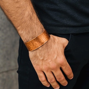 May include: A copper cuff bracelet with a textured, hammered finish. The wide band bracelet is a warm, reddish-brown color and is worn on a wrist. The person is wearing a black shirt and black pants.