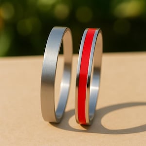 May include: Two silver rings, one with a brushed metal finish and the other with a bright red stripe. The rings are presented on a neutral surface, with a blurred green backdrop.