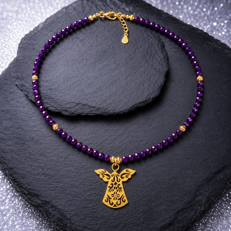 May include: A necklace with small, faceted purple beads and gold details. A gold angel pendant is suspended from the centre. The necklace is presented on a dark, textured surface.