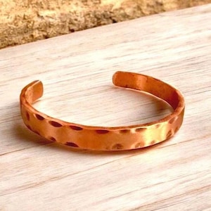May include: A hammered copper cuff bracelet. The open-ended bracelet has a textured surface with small, evenly spaced indentations. The copper has a warm, reddish-brown hue, and the bracelet is displayed on a light-colored wooden surface.