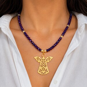 May include: A gold angel pendant necklace with a purple beaded chain. The pendant has a detailed cutout design. The necklace is worn against a white shirt.