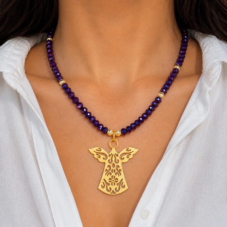 May include: A gold angel pendant necklace with a purple beaded chain. The pendant has a detailed cutout design. The necklace is worn against a white shirt.