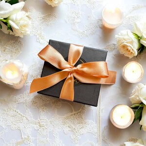 May include: A black gift box with a peach satin ribbon bow. White roses and lit candles are arranged around the box on a white lace surface, creating a romantic and elegant aesthetic.