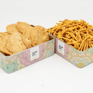 May include: Two colourful tins filled with Indian snacks. The tin on the left contains crispy fried dough, while the tin on the right contains thin, crunchy, yellow sticks. Both tins have a floral design and the text "MUMBAI" printed on them.