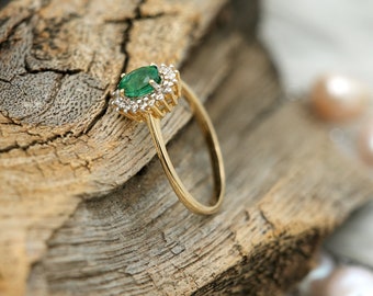 May include: A gold ring featuring a central oval-cut emerald surrounded by a halo of small diamonds. The ring is set against a textured wooden surface, with the focus on the gemstone and the ring's design. The band is thin and polished.