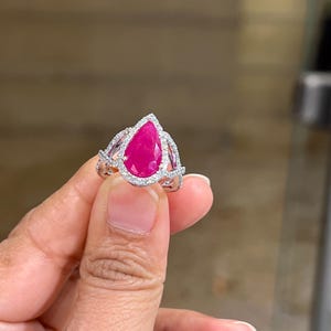 May include: A silver ring featuring a large, pear-shaped, vibrant pink gemstone, surrounded by a halo of smaller, clear stones. The ring's band has an intricate, openwork design, adding to its elegant appearance.