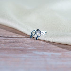 Puede incluir: Un par de pendientes de plata, cada uno con un diseño de símbolo de género. Los pendientes se muestran sobre una tela beige claro y una superficie de madera.